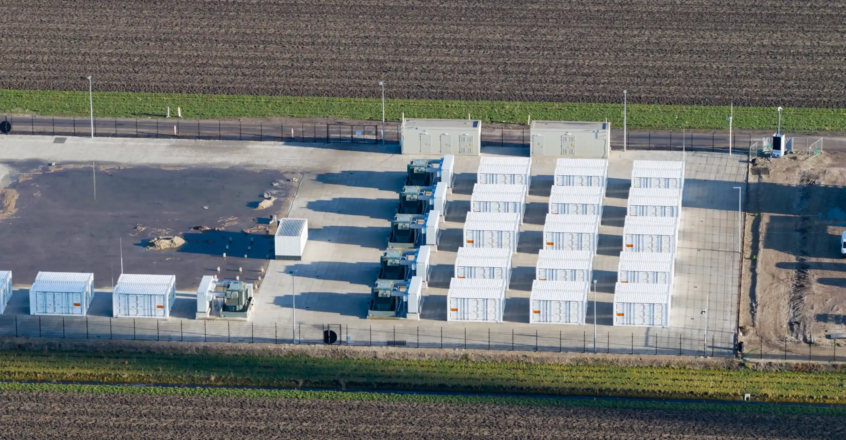 Aerial view of a battery storage compound: rows of white container units on a concrete pad, surrounded by ploughed fields.