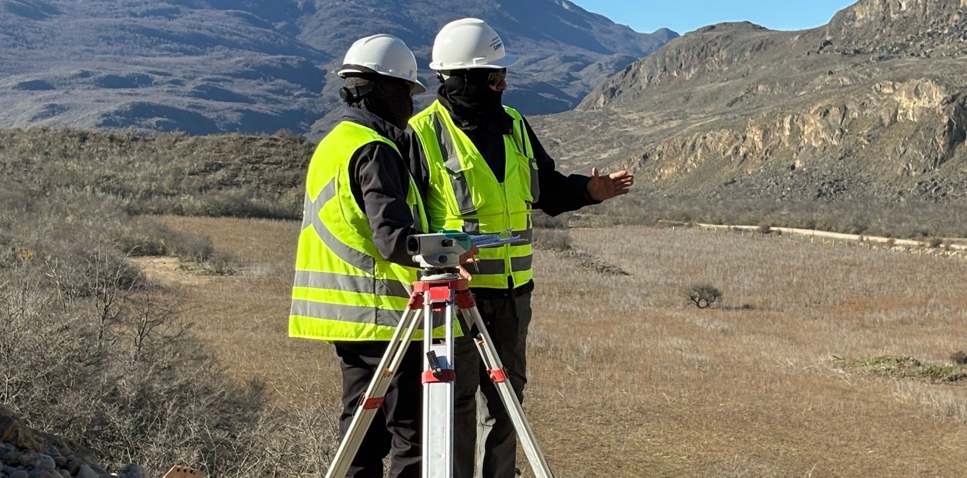 Two surveyors in hard hats and high-visibility vests working with a tripod-mounted instrument, one pointing across an arid valley.